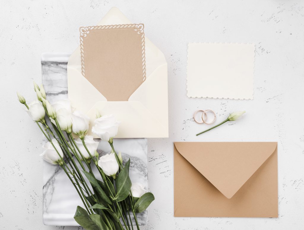 Minimalistyczne zaproszenia ślubne w kopertach kraftowych z białymi kwiatami i obrączkami na tle marmuru.