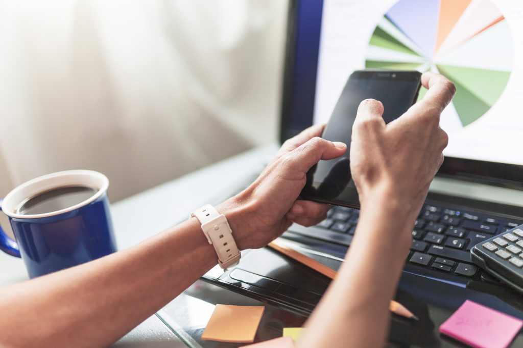 Zbliżenie na dłonie osoby korzystającej ze smartfona przy biurku, w tle widoczny laptop z wyświetlonym wykresem kołowym oraz kubek kawy, symbolizujące profesjonalne płatne reklamy i ich raportowanie.