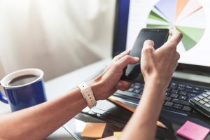 Zbliżenie na dłonie osoby korzystającej ze smartfona przy biurku, w tle widoczny laptop z wyświetlonym wykresem kołowym oraz kubek kawy, symbolizujące profesjonalne płatne reklamy i ich raportowanie.