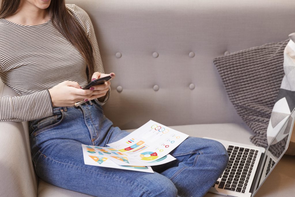 Kobieta siedzi na szarej kanapie i obsługuje smartfon, trzymając na kolanach kolorowe wykresy analityczne, co obrazuje sprawdzanie jak działają płatne reklamy w kampanii marketingowej.